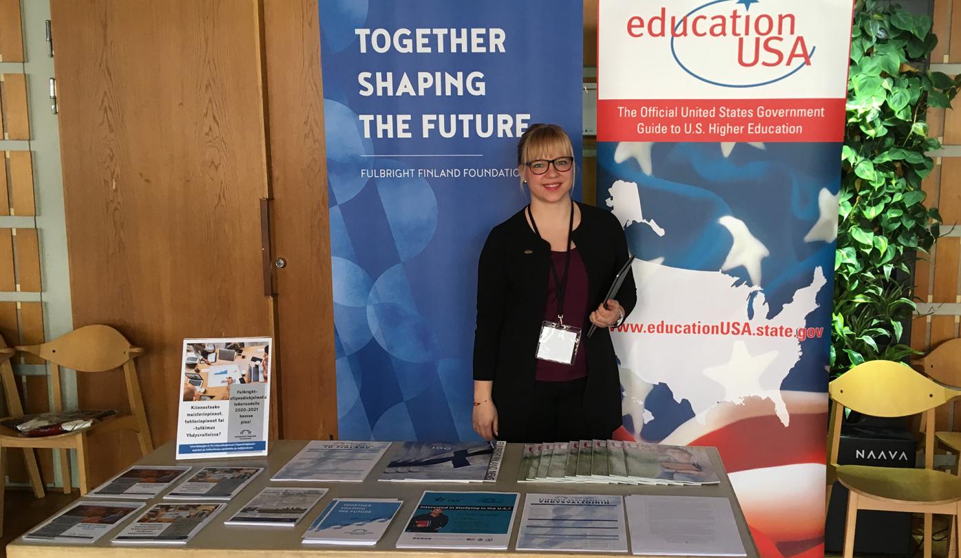 Program Assistant Inari Ahokas at Hanken Exchange Fair, standing behind a table with flyers. There are two roll-ups behind her, one blue with Fulbright Finland Foundation's motto "Together Shaping the Future" and one with EducationUSA logo.