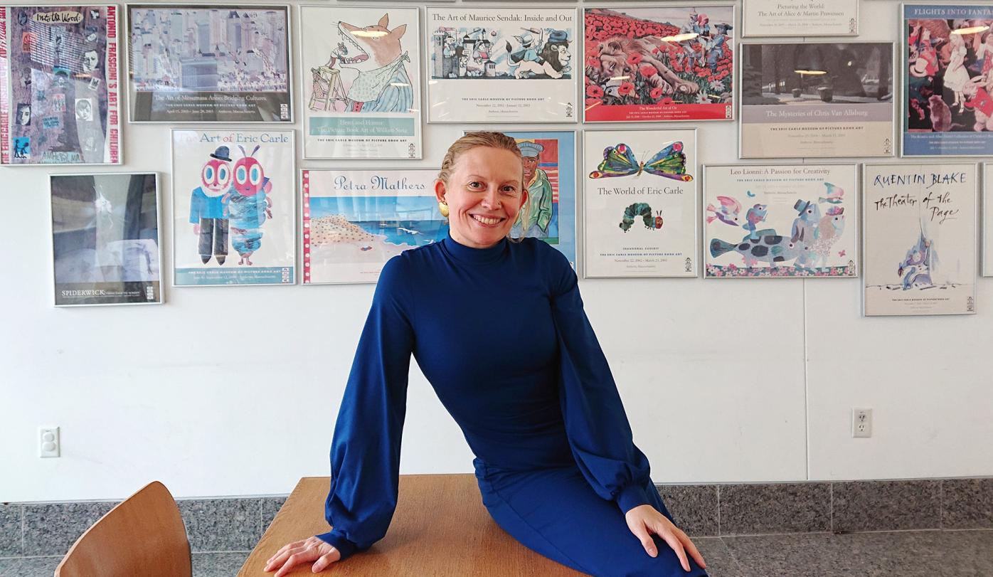 Pirita Tolvanen leaning on a desk, wearing a dark blue dress. She is smiling at the camera. Behind her there are framed illustrations on the wall. 