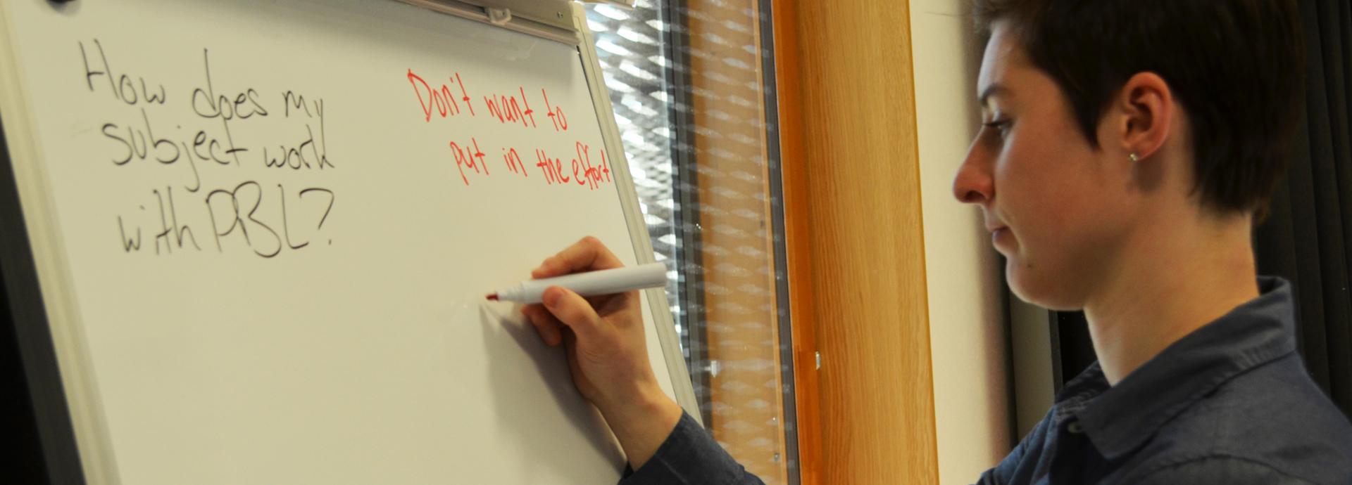 A student writing with a red marker on a white board with questions on it