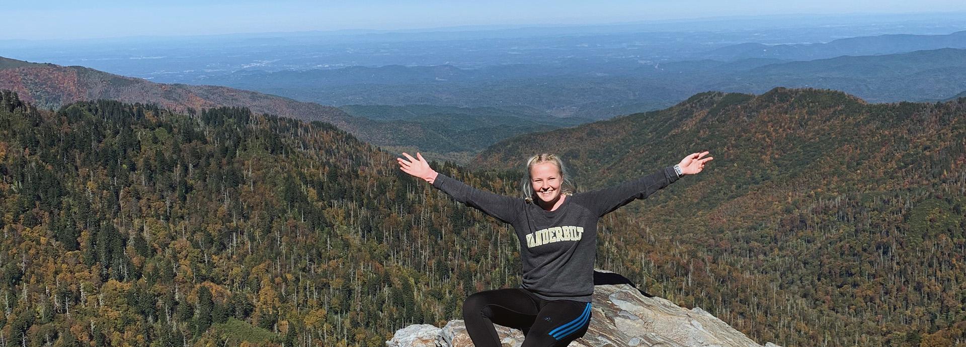 Fulbright Finland grantee Agneetta Moisio sitting on top of a mountain with her arms spread on her side. She is wearing her university's, Vanderbilt University, college shirt and smiling to the camera.