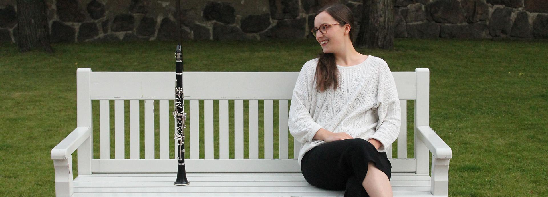 Iines Kiuru sitting on a white bench with a clarinet on her left. She is looking at the clarinet, laughing. Iines is wearing black trousers and white cardigan. 