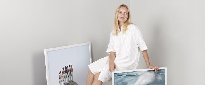Lotta Lemetti sitting and smiling among her photos. She is wearing a white dress and is in a white room.