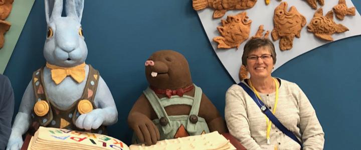 Fulbright Specialist Martha Elford sitting and smiling in front of a dark blue background. There are two large animal characters next to her: a light blue rabbit with a yellow bow tie and brown mole with a red bow tie and green overalls. The animals are reading a big story book.