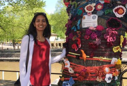 Avanti Chajed standing next to a tree, which is covered in knitted patterns. The photo is taken on a sunny day next to River Aura in Turku, Finland.