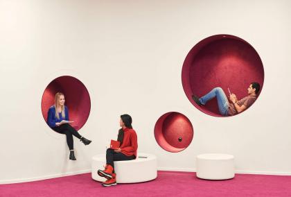 Three students hanging out in a common area where there are red circular holes in a white wall. One female student is sitting in one of the holes and talking with another student in front of her who is sitting on a low white bench. A male student is laying in one of the holes, reading a book.