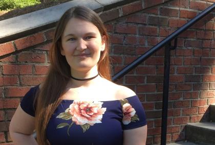 A full body photo of Johna, a young white female with red hair, standing at the landing of an outdoor staircase. She smiles, with one hand placed on her hip. She is wearing a navy blue dress with pink roses.
