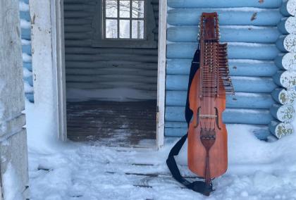 Nyckelharpa leaning against light blue wall of a Finnish wooden house. It's winter, and there is snow on the porch. The door next to the nyckelharpa is open