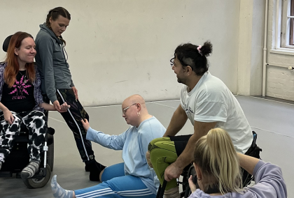 A photographer captures one dancer leaning against another in a wheelchair and one more kneeling on the ground while another wheelchair dancer moves behind.