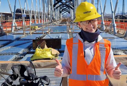 Lada Sokolova on a bridge construction site wearing a orange west, yellow hard hat and giving thumbs up to the camera
