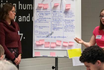 A person explaining what is written on a whiteboard next to them. The whiteboard includes colorful post-it notes and text.