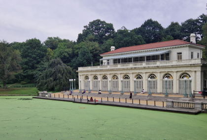 Photograph of a classical-style boathouse with arched windows and a red-tiled roof situated beside a pond covered in green algae. Surrounding dense trees and overcast sky create a serene park setting, with a few people visible on the boathouse terrace.