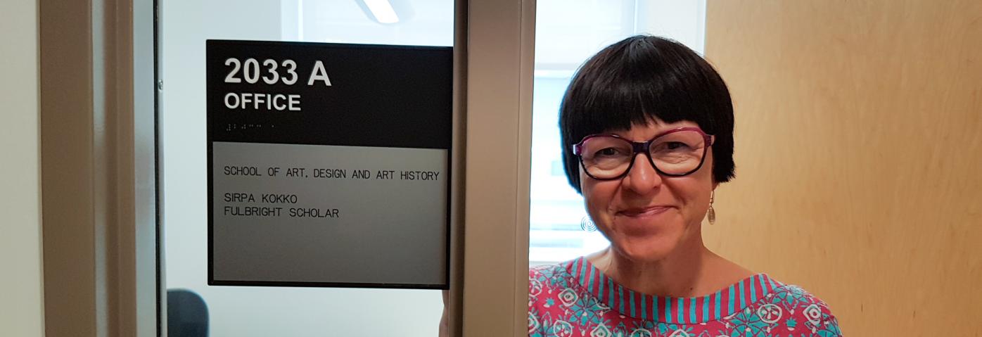 MCPD Sirpa Kokko smiling next to a sign saying "Sirpa Kokko, School of Art, Design, and Art History, Fulbright Scholar"