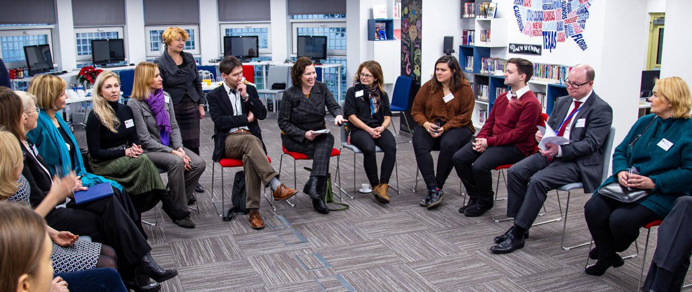 People sitting in a circle. One woman is explaining something with a smile on her face while others are listening to her.