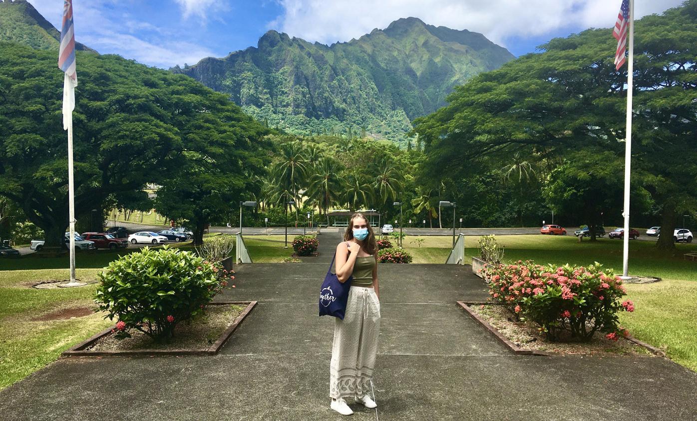 Fulbright Finland Undergraduate grantee Isa Lassinaro at Hawaii Pacific University campus. She is wearing a mask and has a dark blue Fulbright Finland Foundation canvas bag on her shoulder. There are green mountains in the background.