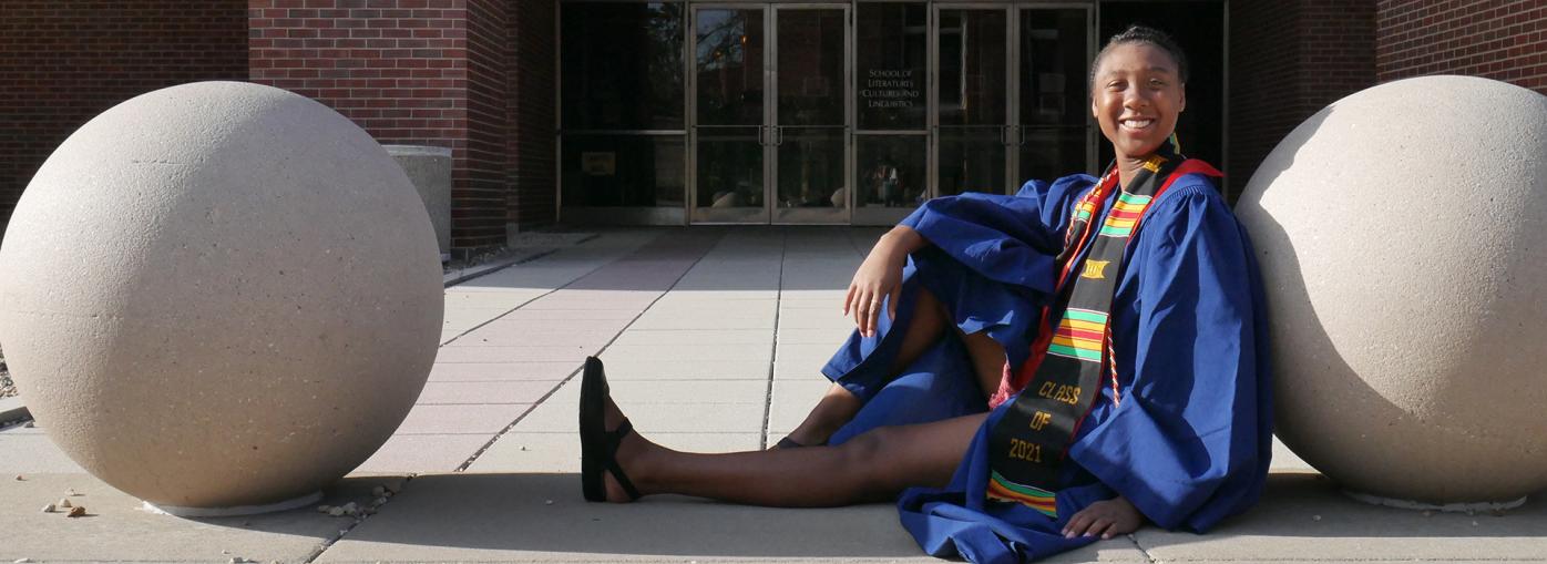 Jade Roberts sitting on a ground, leaning slightly to a concrete sphere, wearing her blue graduate robes. She is smiling widely to the camera.
