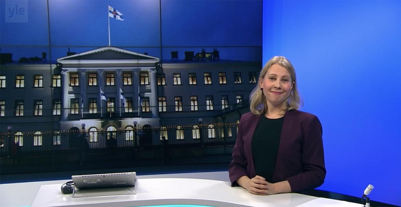 Journalist Kaarina Huovinen standing behind YLE News anchor's table during the news. There is a photo of the Presidential Palace in Helsinki on the background.