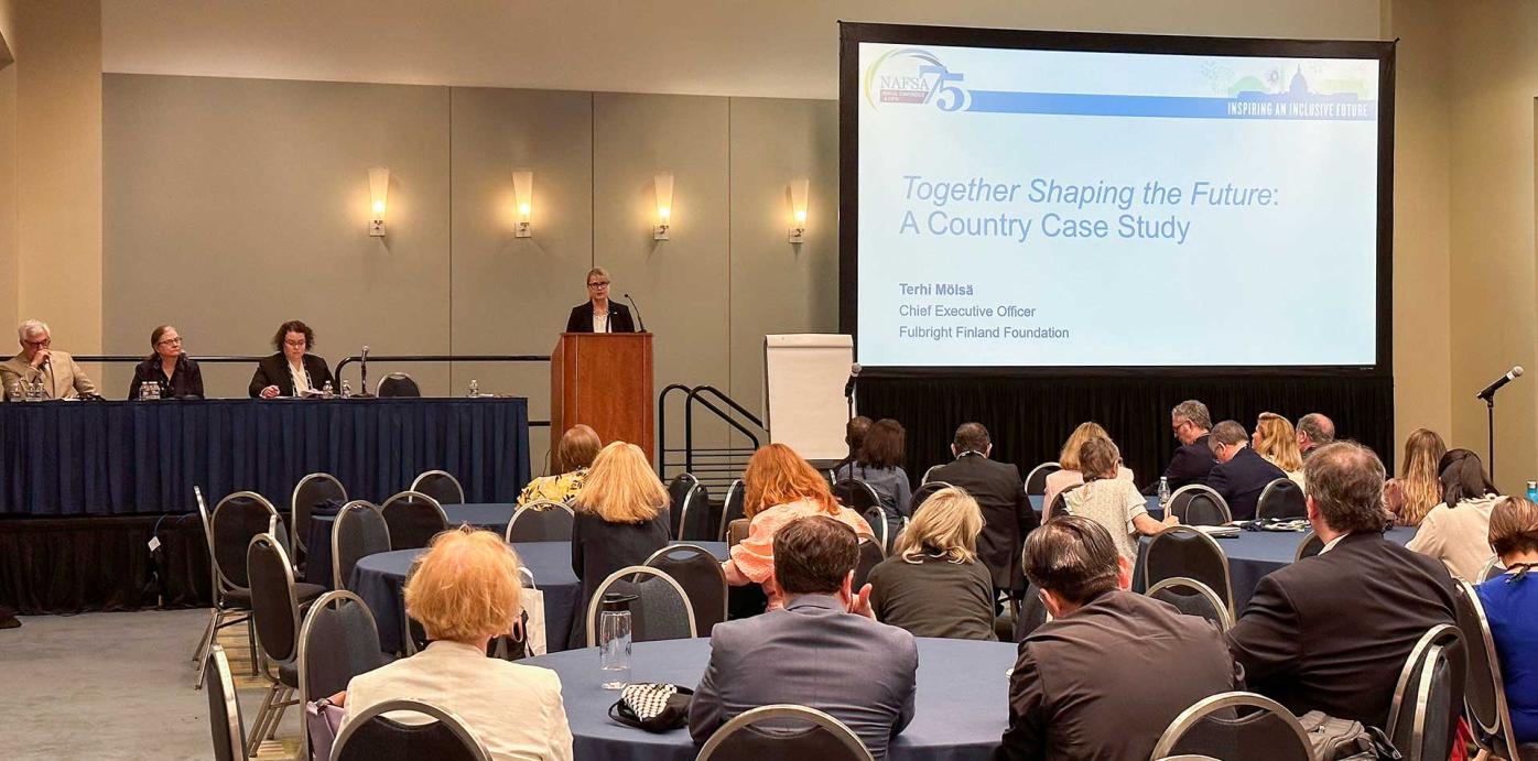 A group of people sitting in chairs at a conference, Fulbright Finland Foundation CEO Terhi Mölsä behind a podium presenting on "Together Shaping the Future: a Country Case Study" at NAFSA 75th conference.