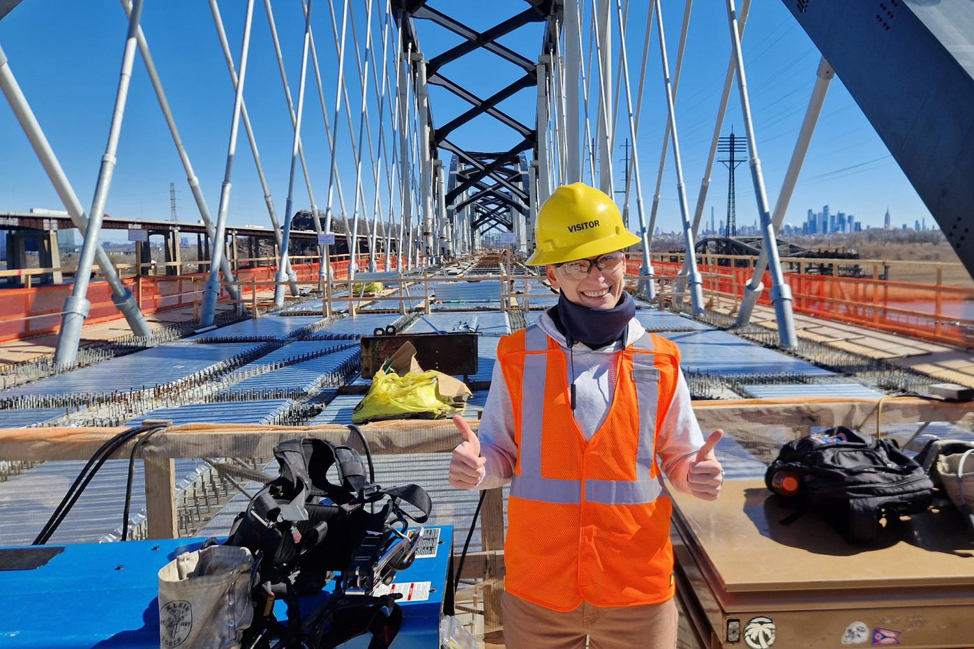 Lada Sokolova on a bridge construction site, wearing orange west and yellow hard hat and giving the thumbs up to the camera