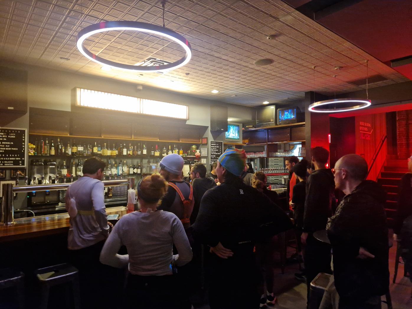Photograph of a dimly lit bar with people in running gear standing and waiting near the counter, illuminated by circular ceiling lights.