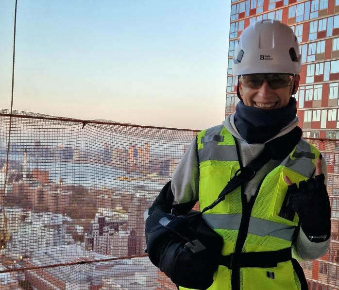 Lada Sokolova on a construction site wearing a hard-hat, safety vest, and a shoulder sling to protect her shoulder. She is smiling at the camera.