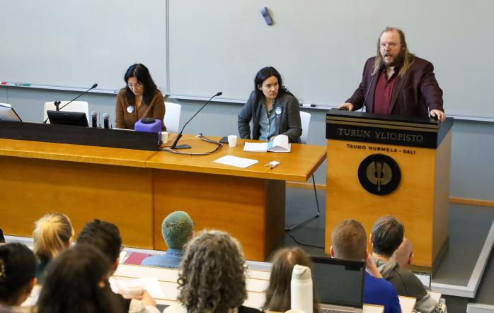 A person talking on a podium, leading a two-person panel discussion at a lecture hall