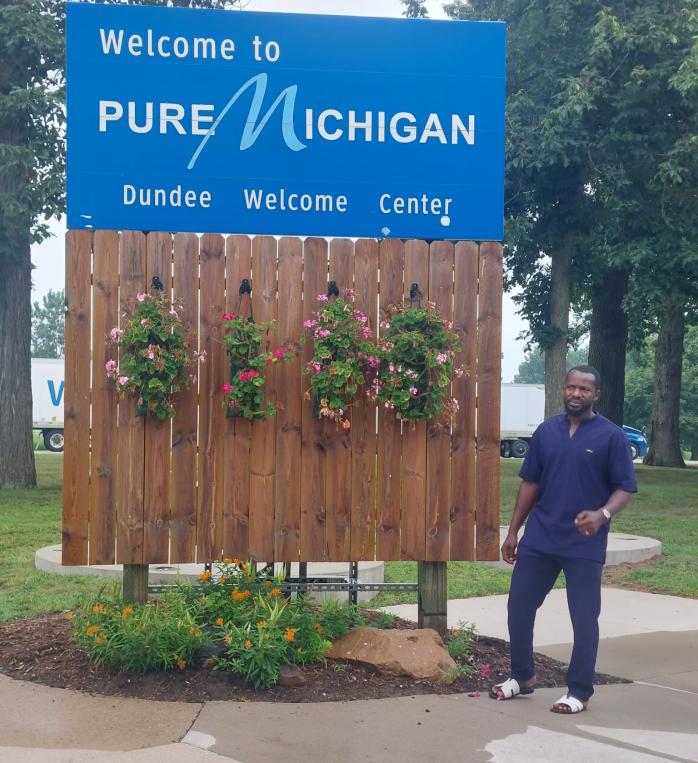 Larry Abdullai in front of a sign saying "Welcome to Pure Michigan Dundee Welcome Center" 