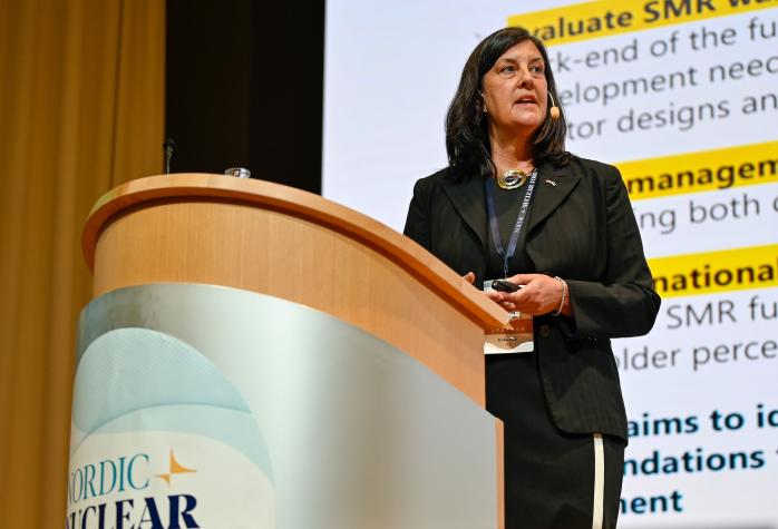 Erika Holt standing behind a podium labeled "Nordic Nuclear" during a presentation. A large screen behind her displays a slide with text.