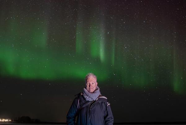 Michael West standing in the middle of the photo with green and red aurora borealis behind him.