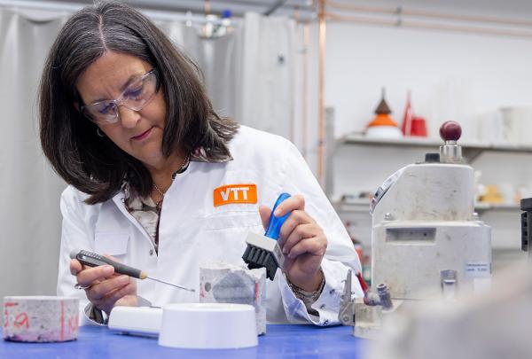 Erika Holt in a white lab coat labeled "VTT" working in a laboratory setting, using a small brush and tool on a white object. Background includes lab equipment, containers, and industrial machinery, indicating a research environment.