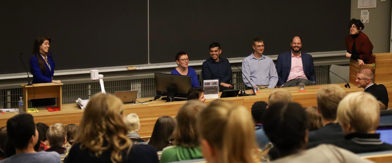 A full auditorium listening to a panel discussion. Five U.S. Fulbrighters are smiling and laughing behind a desk with a moderator smiling at them left of them.