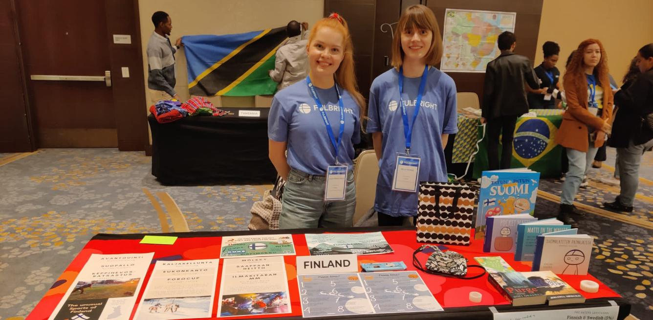 FLTA grantees Laura and Milla wearing Fulbright t-shirts and standing behind a table. The table has red table cloth over it an brochures about Finland.