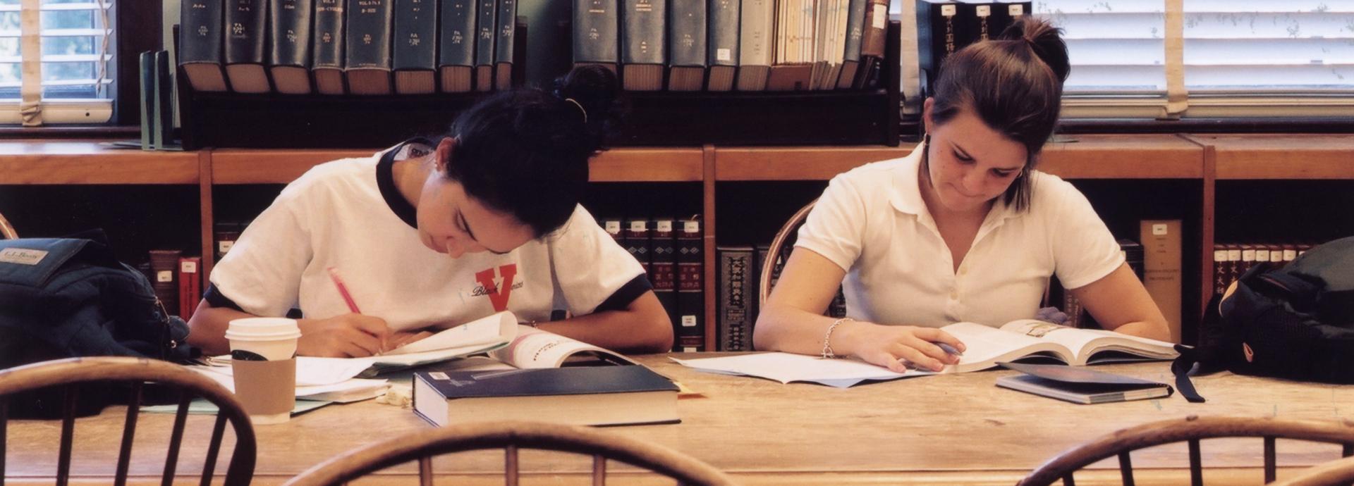 Two female students reading books and taking notes in a library