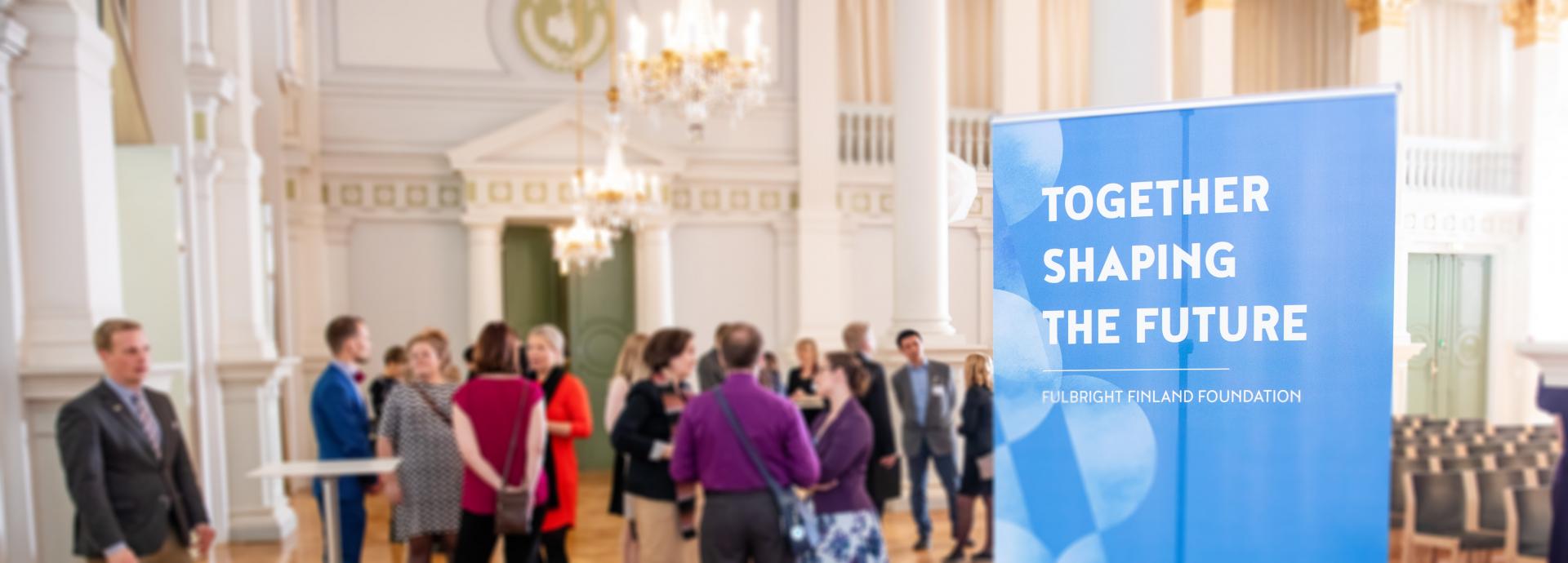 Roll-up with text "Together Shaping the Future" on the front, with people in the background. The photo is from the Helsinki City Hall.