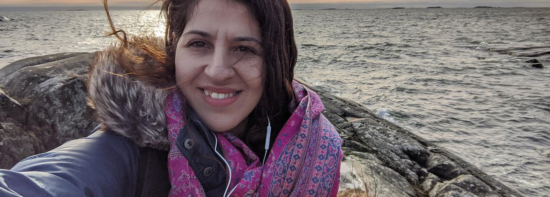 Maedeh Pourrabi on an rocky island, taking a selfie on a windy evening with sea and sunset behind her.