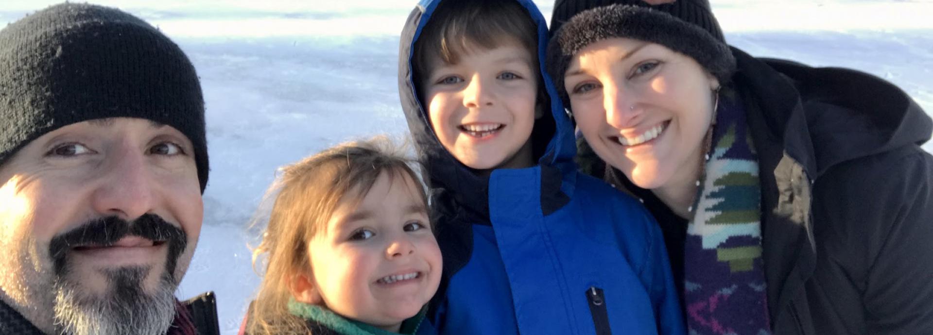 Family of four in a winter clothes smiling widely to a camera. The father on the left is taking a selfie of them, and they are standing on a frozen lake on a sunny day.