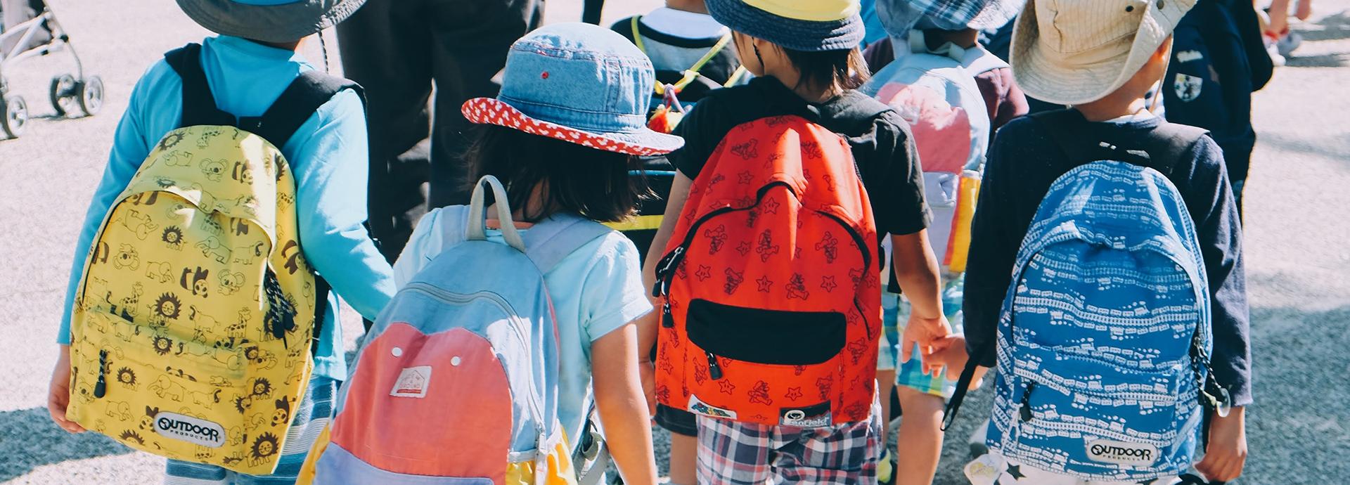 A photo of four little kids on their way to school. They are wearing colorful backpacks.