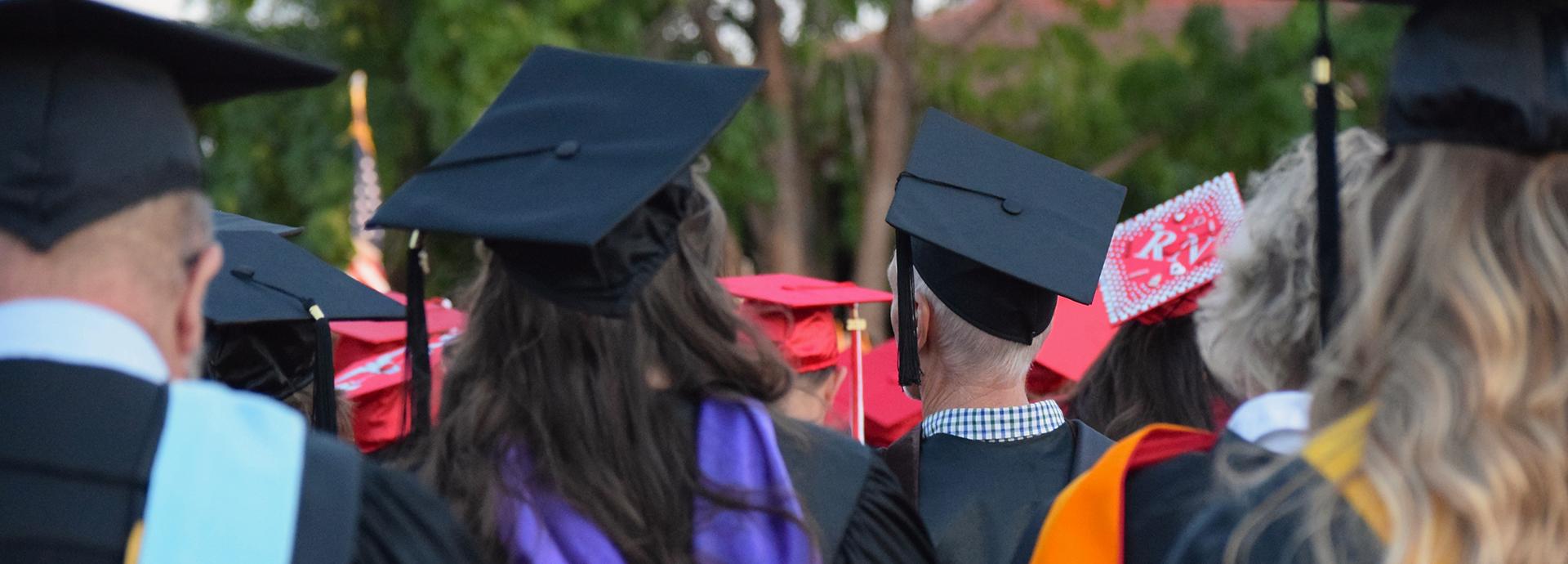 Opiskelijoita graduation-hatut päässä, kuva otettu yleisön takaa.