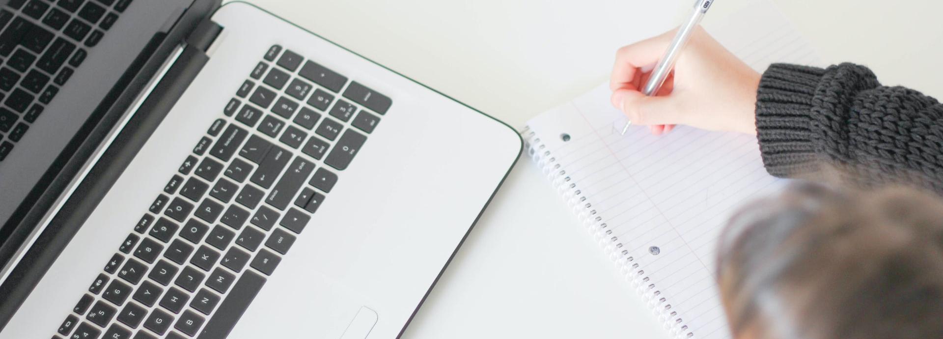 There is a corner of a laptop on the left and a person writing on a blank piece of notebook paper