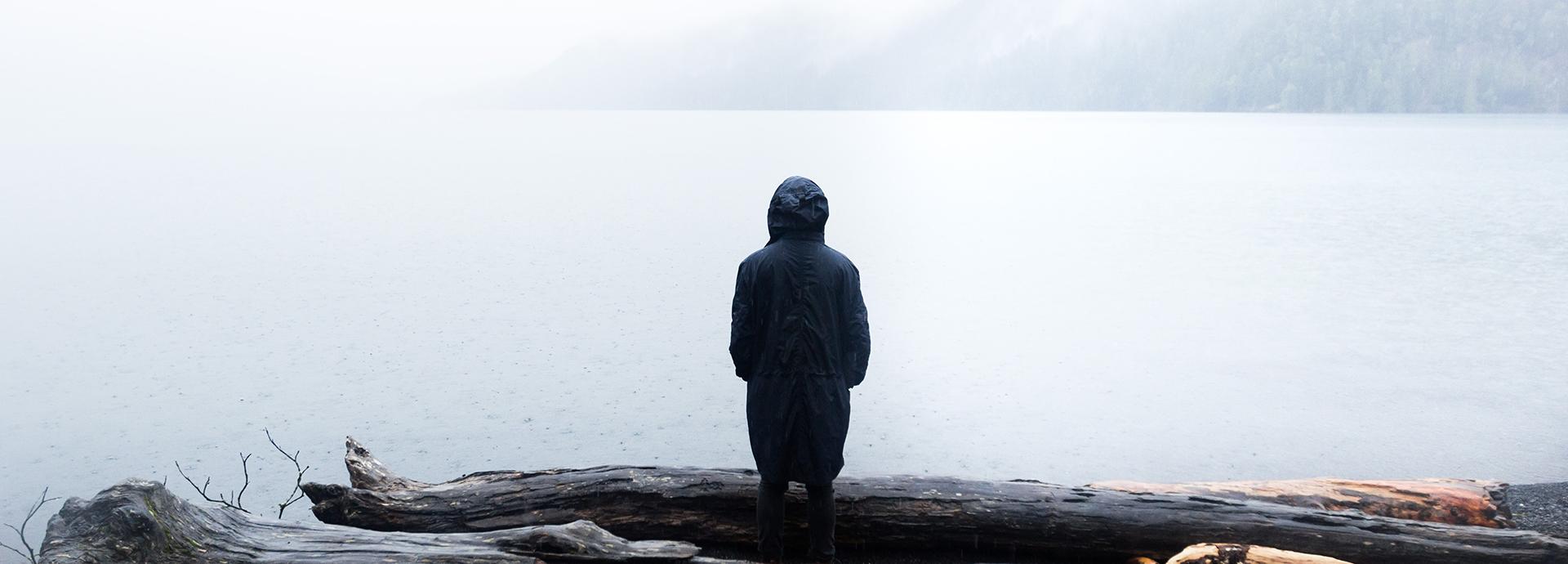 A person wearing dark coat standing by themselves on a shore of a misty lake, looking to the lake and standing their back to the camera.