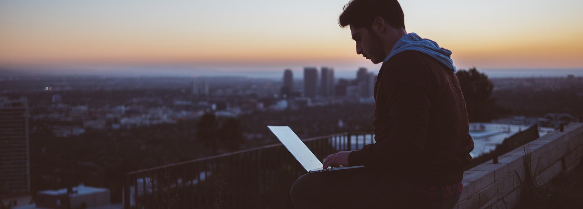 A man looking at a laptop screen, and sitting on a wall with a city skyline on the background