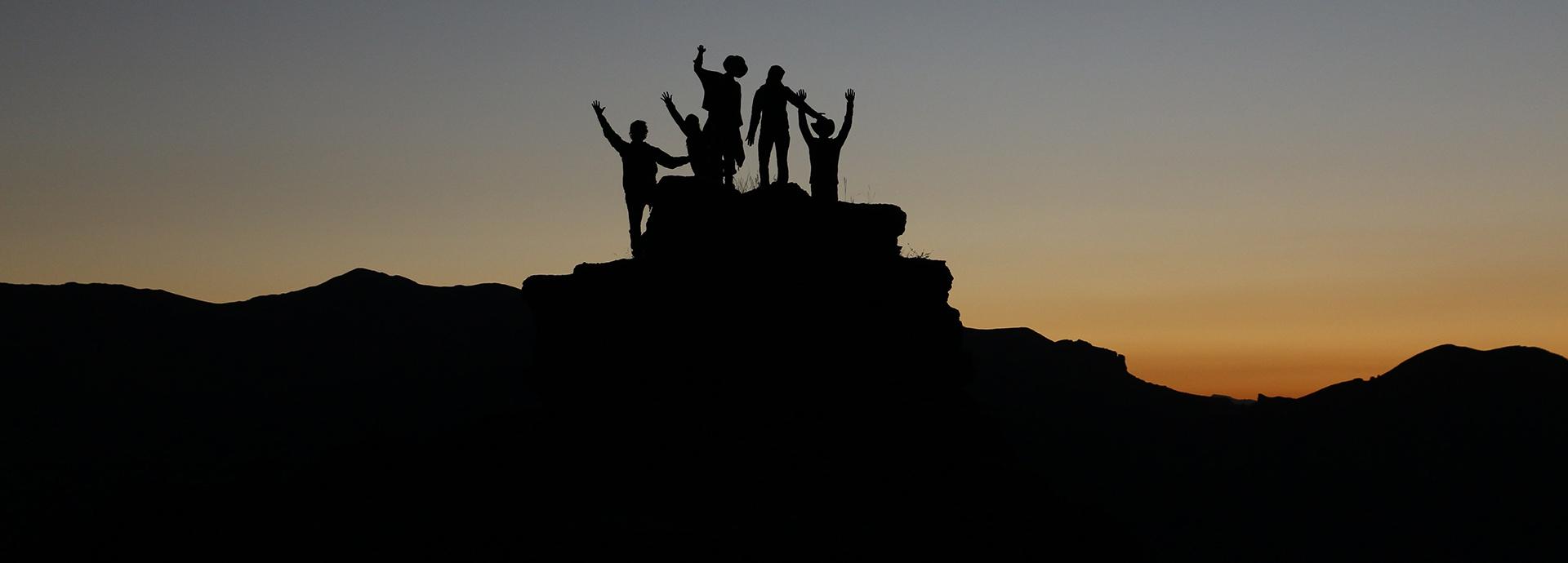 Silhouette of group of five people on a mountain during a sunset. The people have their hands up.