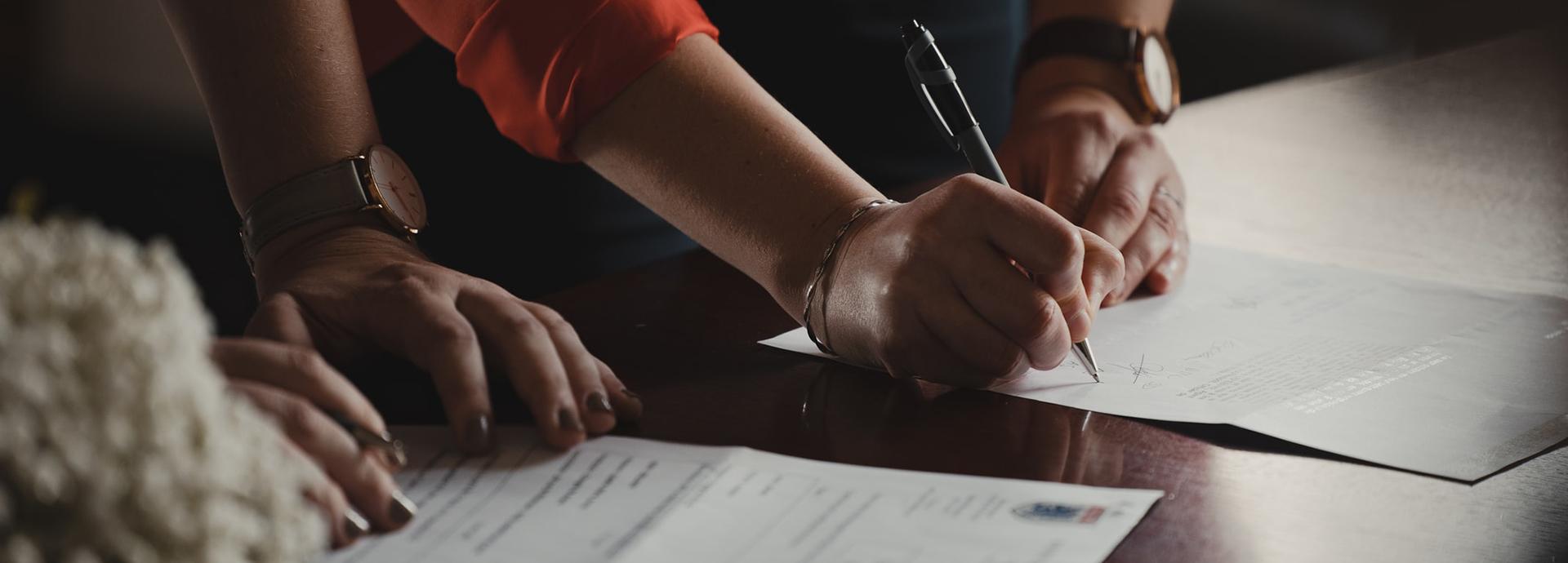 There are documents on a table and someone signing one of them with a pen. There is another person next to the one who signs the paper, their hands on the table.