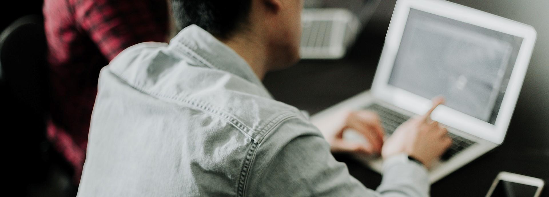 A person wearing a light blue shirt, working with a laptop, the photo has been taken behind their shoulder.