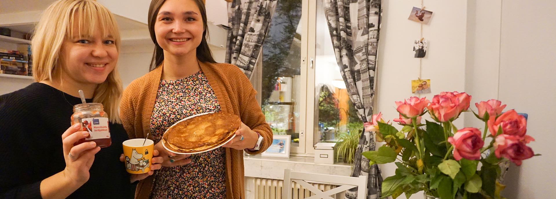 Fulbright alumna and a grantee smiling at the camera. They are holding a plate of Finnish pancakes they have just made and a jar of jam. There are pink roses on in a vase on their right.