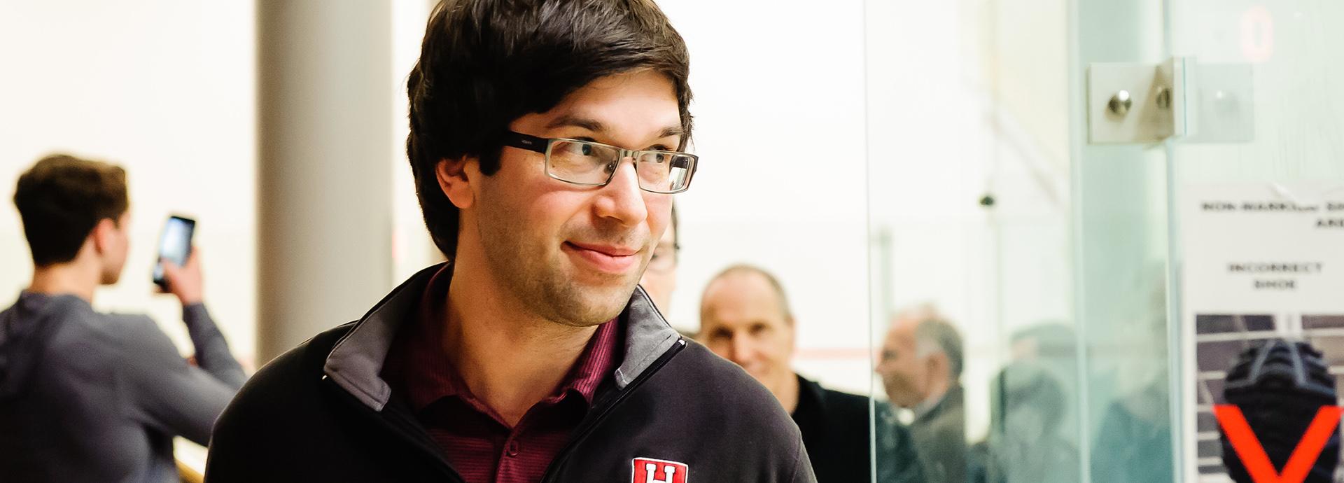 Hameed Ahmed is looking to his left with a light smile. The photo is taken in a squash hall.