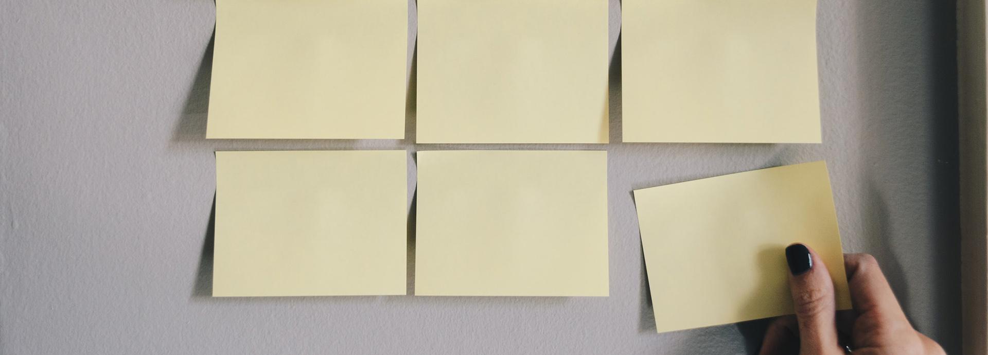 Six light yellow sticky notes on a wall. The note in the lower right-hand corner is being pulled out by a hand that has black nail polish.