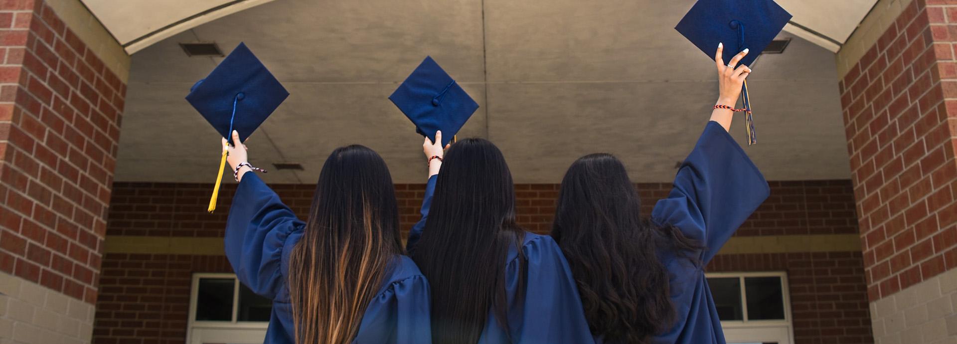 Three female students facing away from the camera in their graduation robes lifting up their graduation caps. 