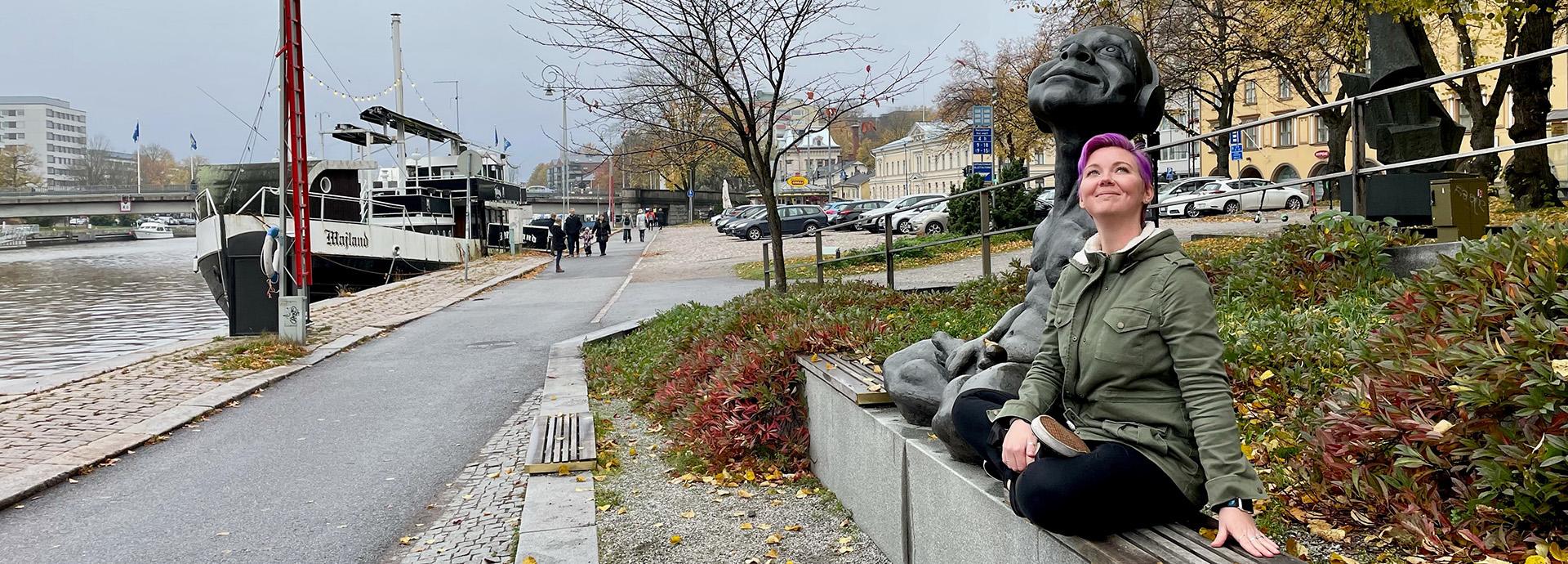 Meg Jones sitting next to statue, looking up with a smile on her face. The statue is on a bench on a river bank in Turku and there is a river boat in the photo on the left.