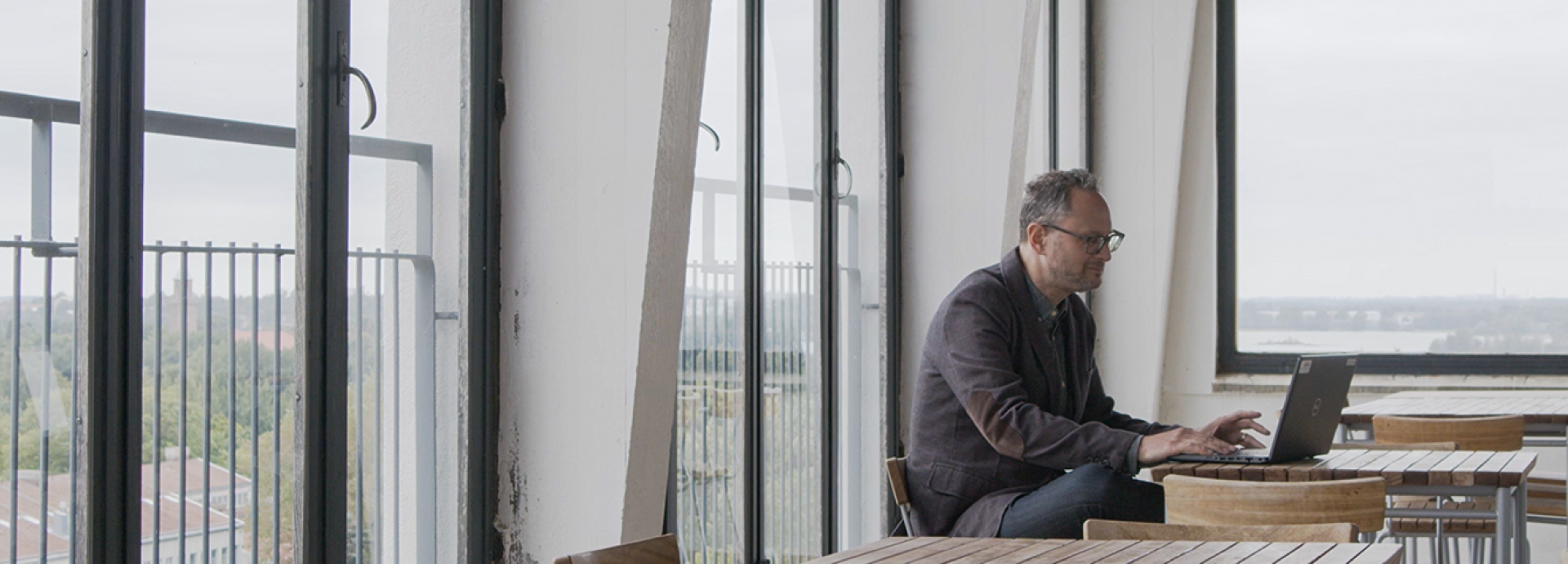 Fred Markowitz sitting, working his laptop which is on a table in front of him. The photo is taken on a high terrace and there are large windows behind him.