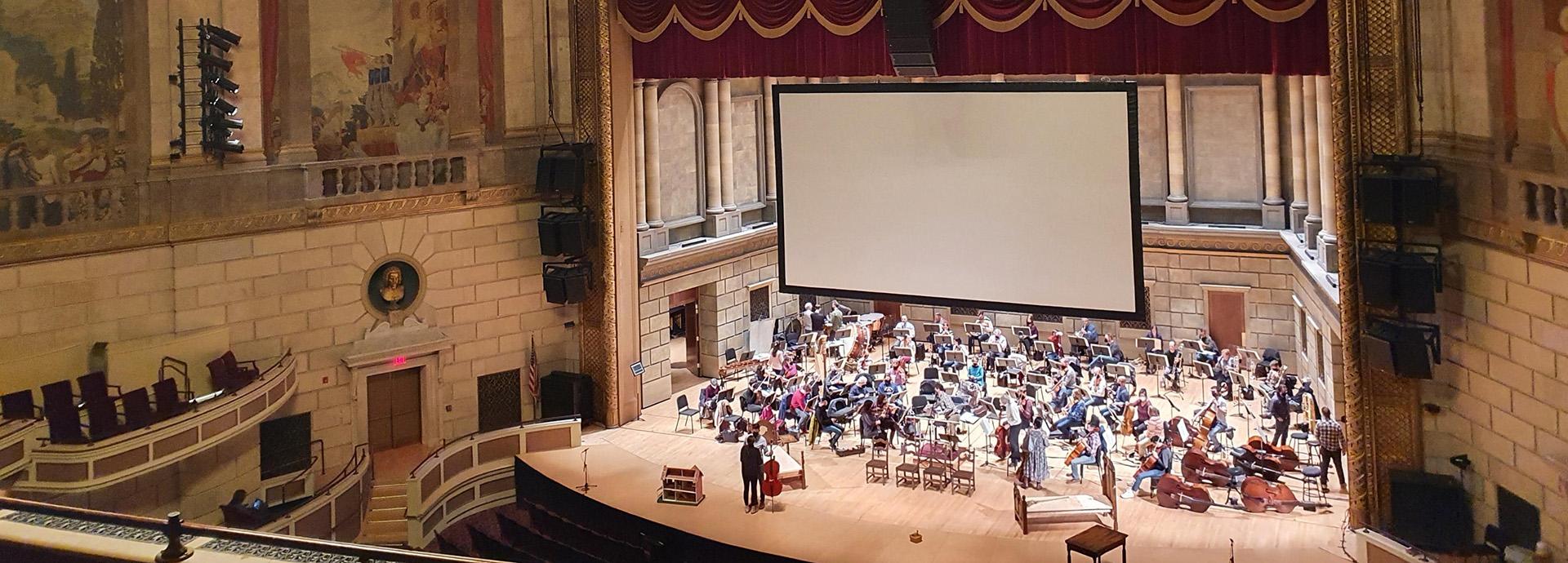 Photo of orchestra on a stage taken from the balcony of a music hall.